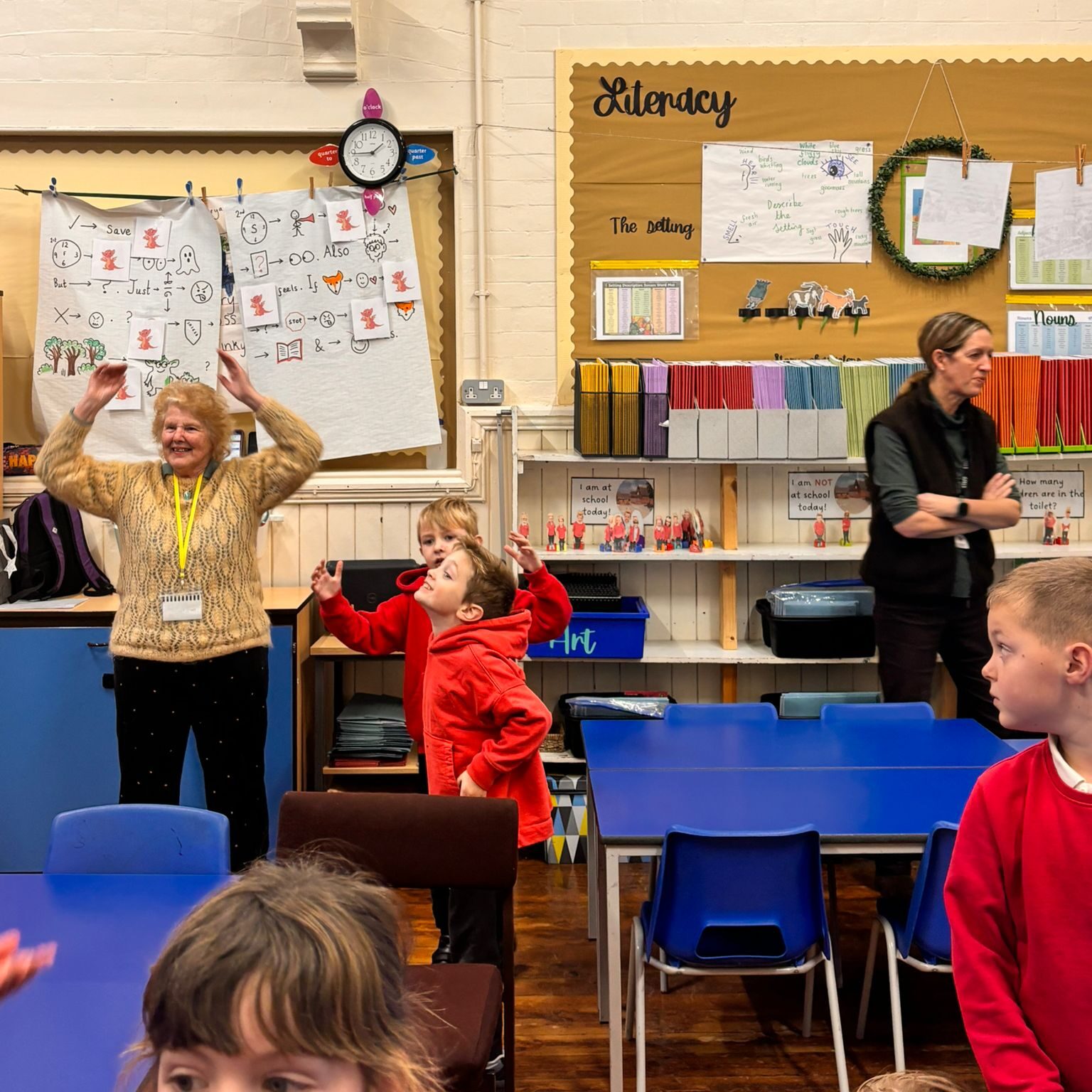 A classroom scene with children and adults. A woman in a yellow sweater is energetically raising her arms, engaging with the children, who are dressed in red uniforms. Another adult in black stands near a bulletin board labeled "Literacy," which has student work and educational materials. The room has colorful storage bins and desks with blue chairs.