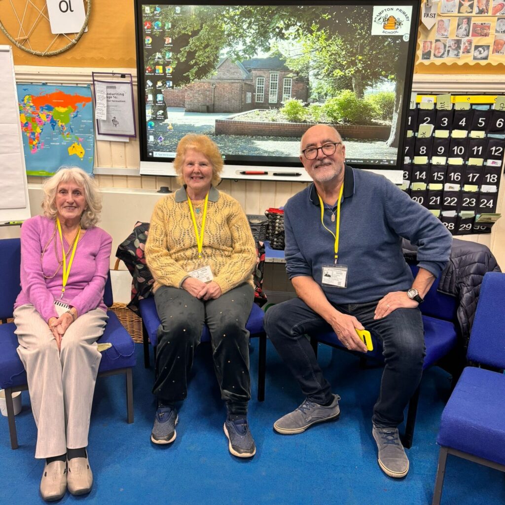 Three older adults sitting on blue chairs in a classroom. The woman in the middle wears a yellow sweater, while the woman on the left is in purple and the man on the right is in a navy-blue sweater, holding a yellow phone. Behind them is a digital screen displaying an image of a school building, a world map, and educational posters.