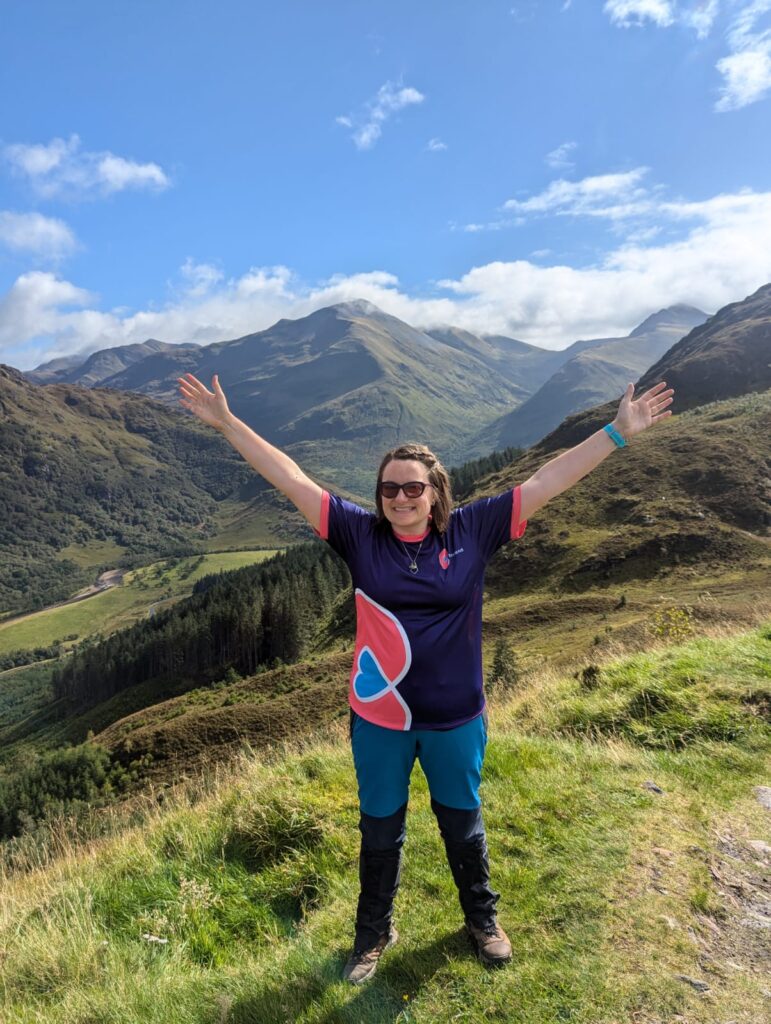 A woman standing in a meadow with montains in the background. She wearing sunglasses and a branded purple b:friend T-shirt, she is smiling and stretching her hand out towards the sky.