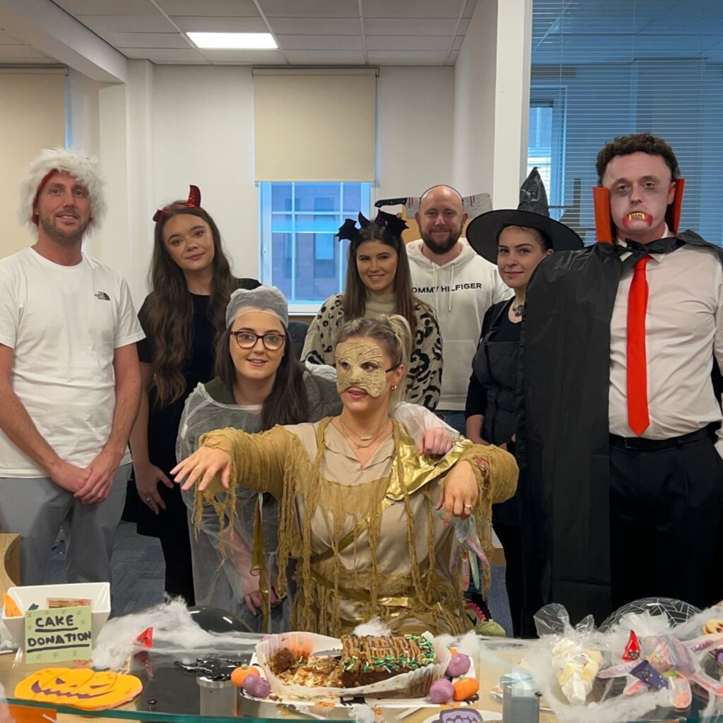 A group of people wearing Halloween costumes in an office with plates of cakes in front of them